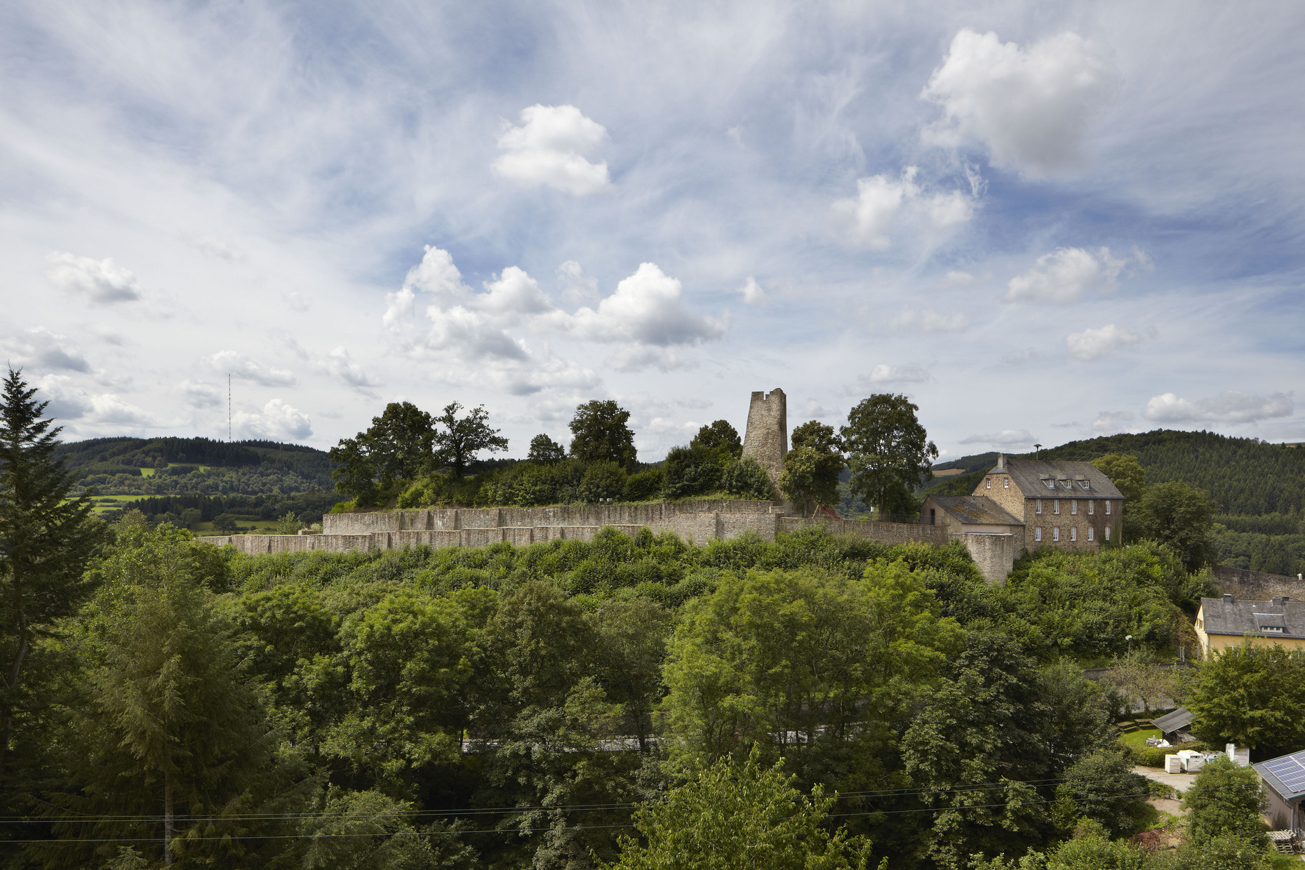 Anhöhe mit Bergfried, Teilen einer Umfassungsmauer und Häusern, bewaldete Hügel im Hintergrund